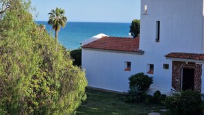 A white building with a red-tiled roof, a small garden, and a view of the sea.