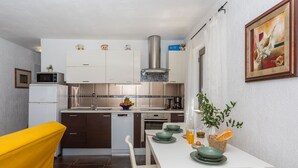 A compact kitchen with white cabinets, a microwave, and a stainless steel range hood. There is a dining table with green plates and a yellow chair. - Apartment Anivija (Omisalj)