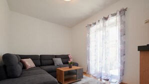 A modern living room with a grey sectional sofa, a wooden coffee table, and a floral-patterned window with sheer curtains. - Cozy Downtown Trogir Apartment With Balcony (Trogir)