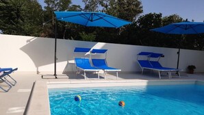 Una piscina con sedie e ombrelloni azzurri, circondata da un muro bianco e alberi.