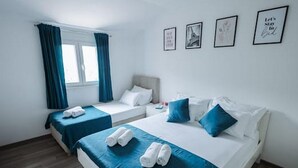 Hotellihuone, jossa on kaksi sänkyä, ikkuna, jossa on verhot, ja neljä kehystettyä kuvaa seinällä.
