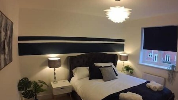 A bedroom with a bed, bedside lamps, a nightstand, a potted plant, and a window with a view. - Modern Home From Home With Cinema - Elm Tree House (Leicester)