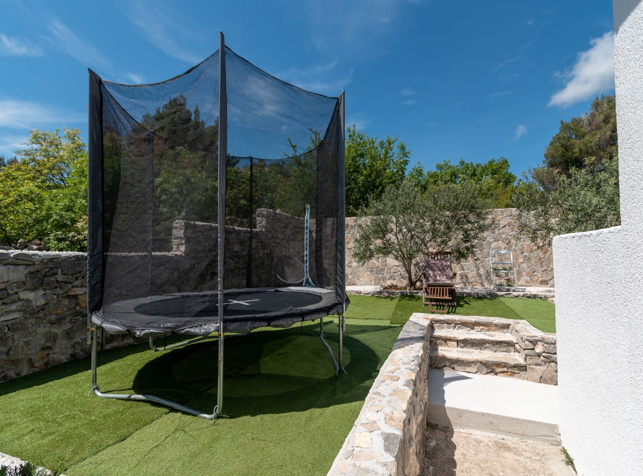 A trampoline with a safety net in a backyard with stone walls and a bench.
