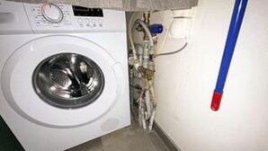 A compact, white washing machine in a small laundry room with a blue and red pipe on the wall. - 4 Star Holiday Home in Hejls (Hejls)