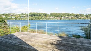 Um deck de madeira com guarda-corpo de metal, com vista para um lago calmo, rodeado por vegetação e casas ao longe.