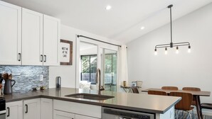 Une cuisine moderne avec des armoires blanches, un lave-vaisselle en acier inoxydable et un grand îlot central doté d’un évier. On y trouve un coin repas avec des chaises et une table en bois, ainsi qu’une vue sur un balcon accessible par une porte vitrée.
