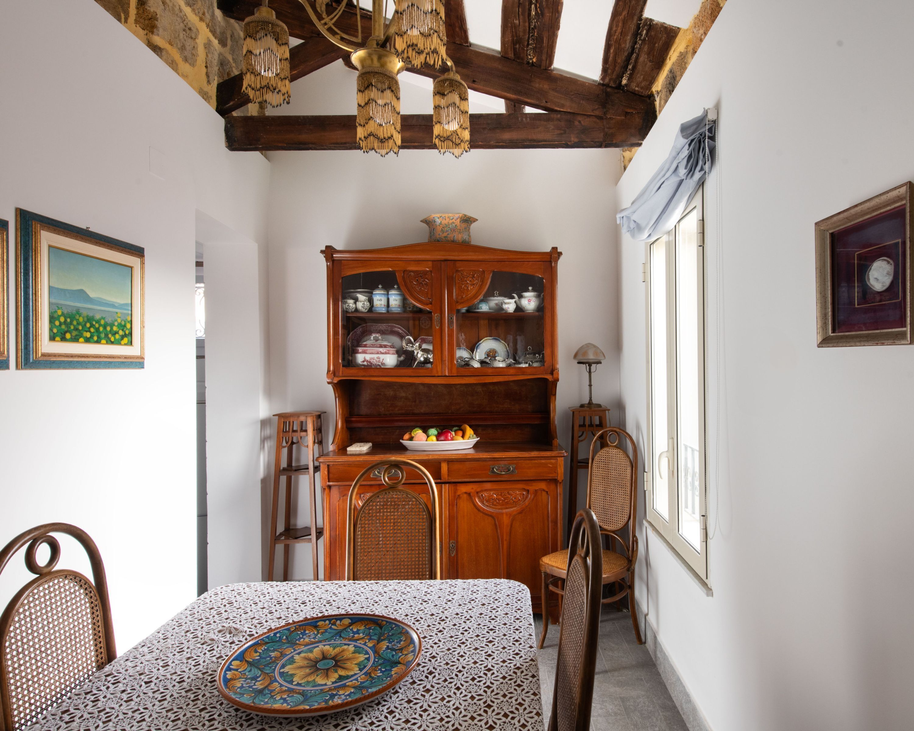 Un comedor con una mesa de madera, sillas, mantel de encaje, un aparador con vajilla y un cuadro en la pared.