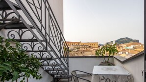 Ένα μπαλκόνι με σιδερένια κάγκελα, ένα τραπέζι με τραπεζομάντιλο δαντέλας και γλάστρες με φυτά.