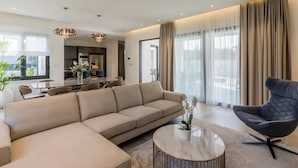 Ein modernes Wohnzimmer mit einem Ecksofa, einem runden Couchtisch und einem schwarzen Sessel. Im Hintergrund ist ein Essbereich mit Tisch und Stühlen erkennbar.
