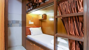 A compact, single-bed room with a wooden headboard, a small bedside table, and a wall-mounted light fixture. - Neon Nest Dormitory Suits (Mumbai)