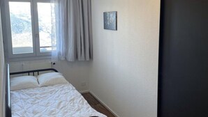 Ein kleines Zimmer mit einem Einzelbett, einem Fenster, einem Vorhang und einem kleinen Bild an der Wand.