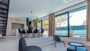 Uma sala de estar moderna com uma janela grande, uma área de jantar com cadeiras pretas e uma cozinha ao fundo.