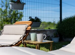 Una mesa de madera con una taza blanca y una taza verde, un banco de madera con un cojín color beige, y una maceta colgante con flores.
