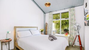 A bedroom with a wooden bed, a bedside table with a lamp, a window with curtains, and a fan. - Pelican Cottage - Pet Friendly - Wifi (Goolwa North)