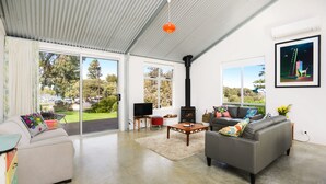 A spacious living room with a fireplace, comfortable seating, and large windows offering a view of the outdoors. - Pelican Cottage - Pet Friendly - Wifi (Goolwa North)