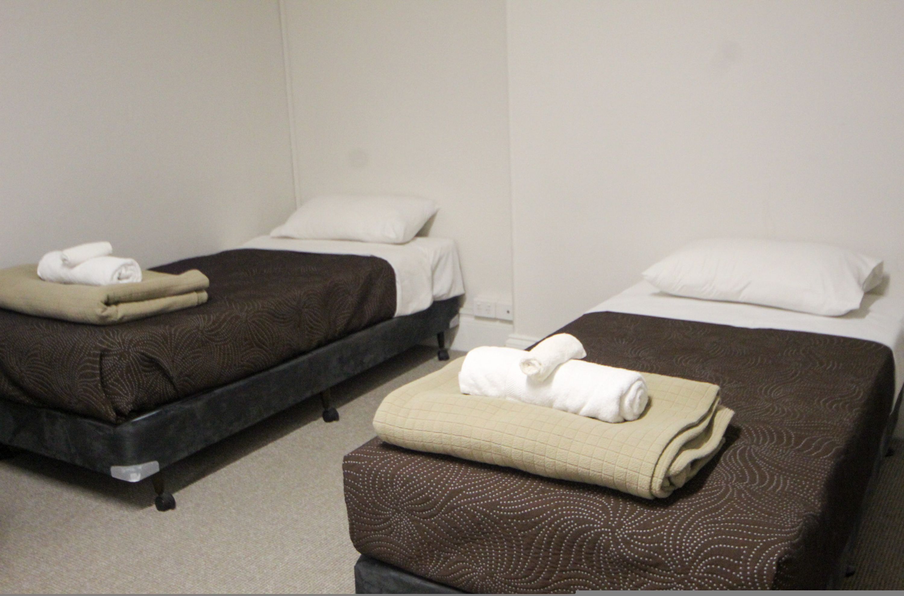 Two single beds with brown bedding and white pillows, each with a rolled towel on top.