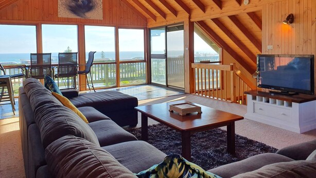 A spacious living room with a large sofa, a coffee table, and a television. There are large windows offering a view of the ocean.