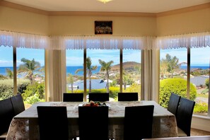 Un comedor con una mesa preparada para cuatro, ventanas amplias con cortinas blancas y vistas al océano y follaje.