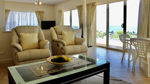 Un salón con dos sillones color beige, una mesa de centro de vidrio con un cuenco de frutas y un televisor en la pared.