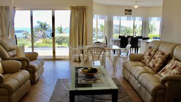 A living room with a glass coffee table, beige sofas, and a dining area with white chairs.