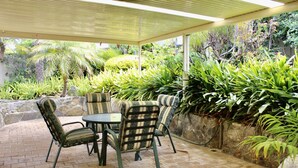 Un patio al aire libre con una mesa y sillas, rodeado de abundante vegetación y un muro de piedra.