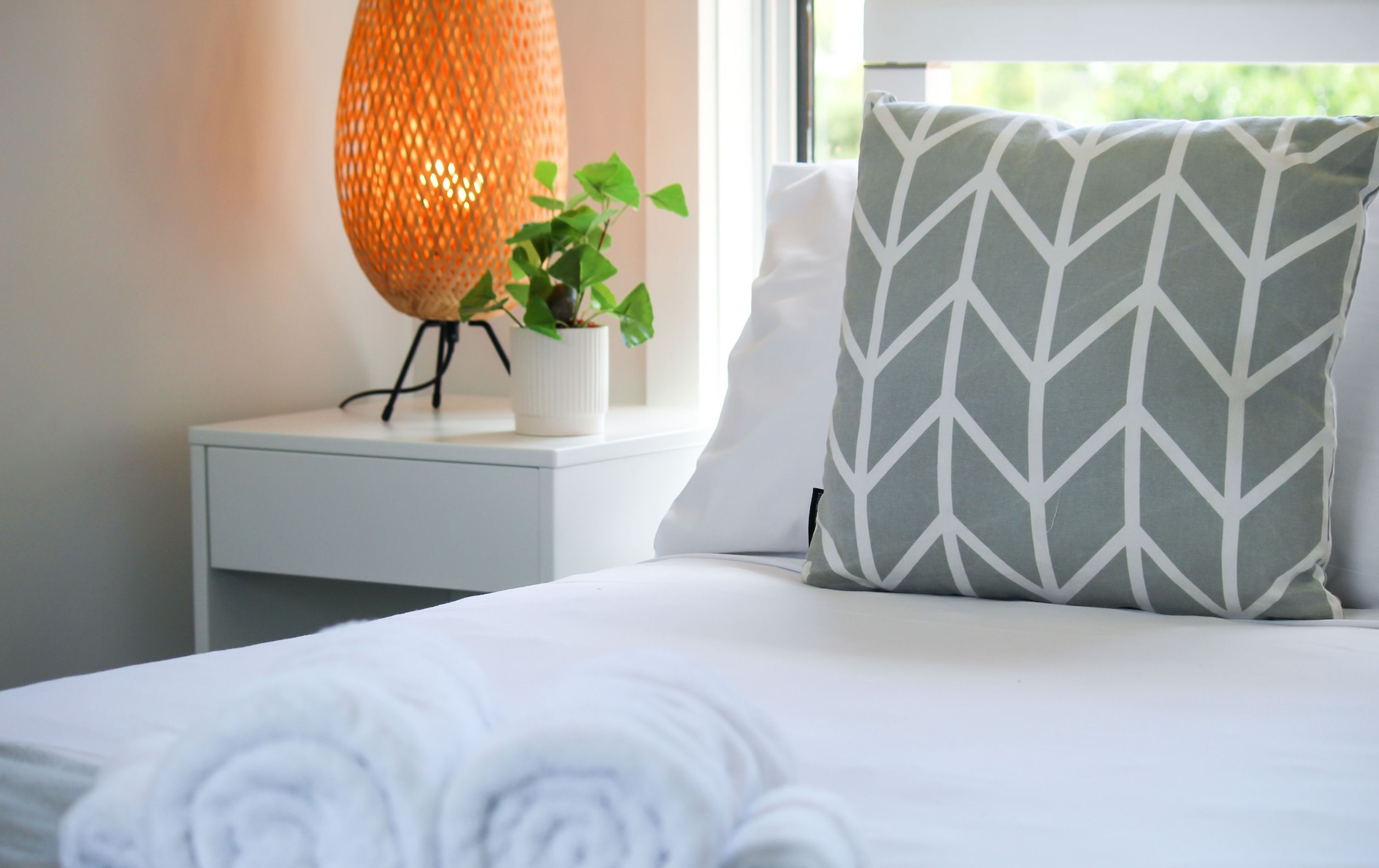 Un lit bien fait, recouvert d’une couette blanche et agrémenté d’un coussin à motifs chevrons gris et blancs. On y trouve une table de chevet blanche, sur laquelle sont posés un pot de plante et une lampe orange à texture particulière.