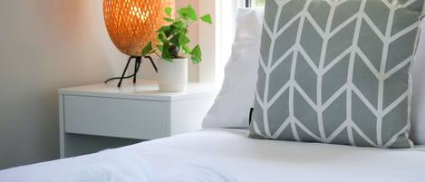 Un lit bien fait, recouvert d’une couette blanche et agrémenté d’un coussin à motifs chevrons gris et blancs. On y trouve une table de chevet blanche, sur laquelle sont posés un pot de plante et une lampe orange à texture particulière.