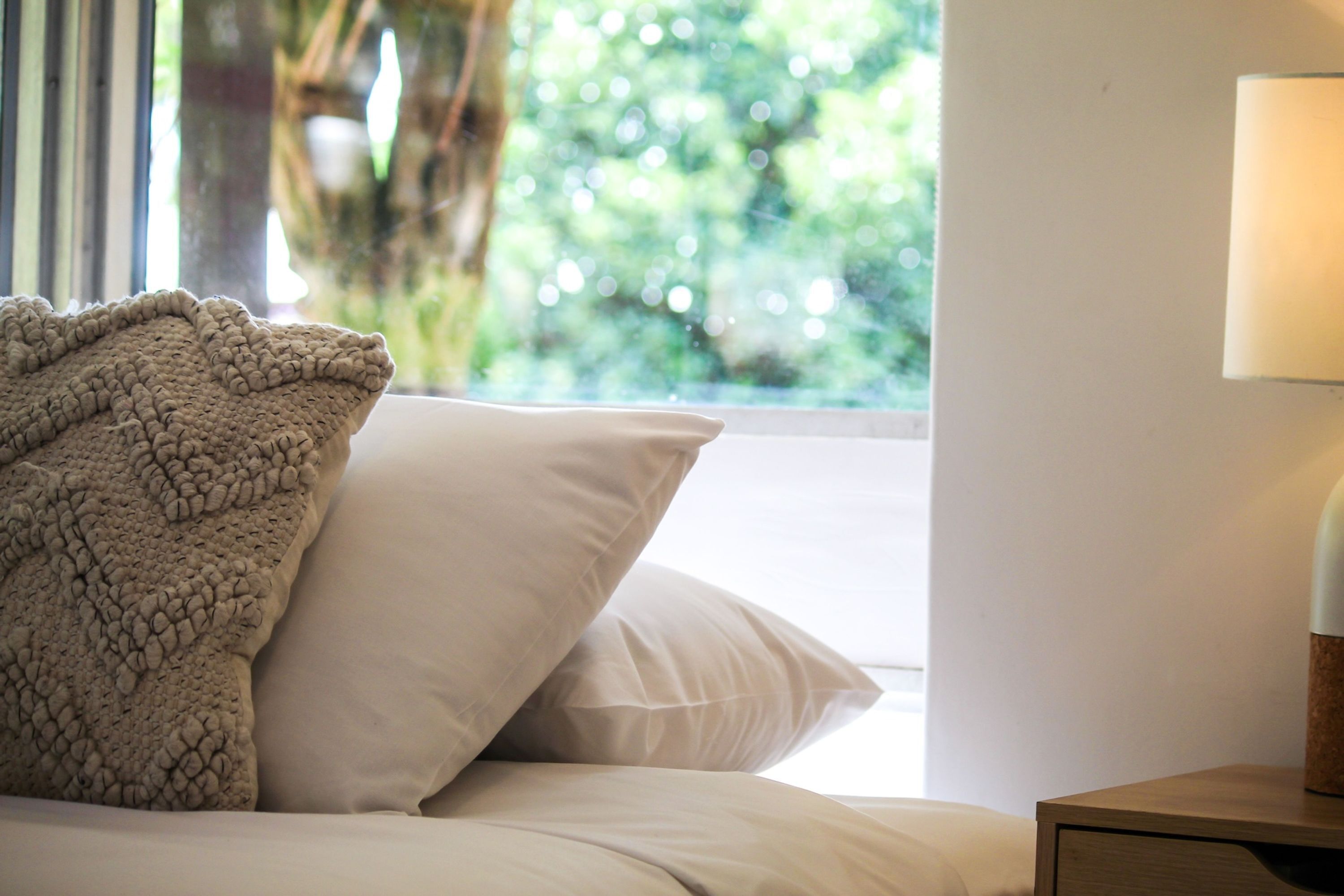 A bed with a textured blanket and pillows, a bedside lamp, and a window with a view of greenery.