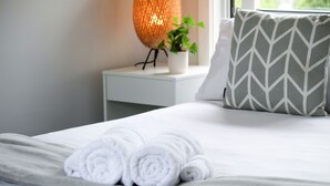 A neatly made bed with rolled white towels, a white nightstand with a potted plant, a patterned pillow, and a textured wall lamp. - Sandy Pool Shack - Pet Friendly (Seacliff Park)