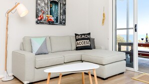 Een moderne woonkamer met een lichtgrijze modulaire bank, een witte salontafel en een vloerlamp. Op de muur hangt een schilderij van een scooter en een deur, en op de bank ligt een kussen met de tekst 'BEACH ACCESS'.