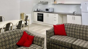 A compact living space with a kitchenette, a dining area, and a seating area with patterned sofas and red cushions.