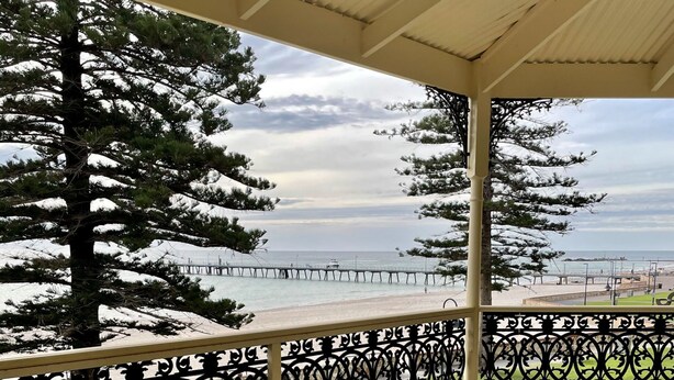 A covered outdoor area with a wooden deck and metal railing, overlooking a beach and a pier.