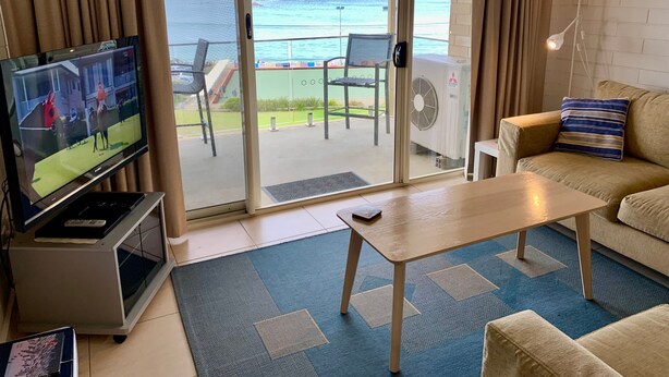 A living room with a television, a sofa, a coffee table, and a view of the ocean through a sliding glass door. - The Dolphins 7 - A View To Remember (Port Elliot)