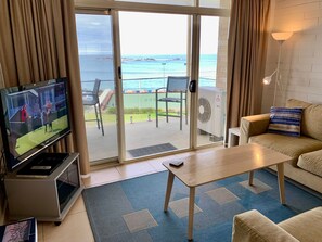 A living room with a television, a sofa, a coffee table, and a view of the ocean through a sliding glass door. - The Dolphins 7 - A View To Remember (Port Elliot)