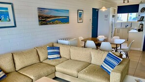 A modern living room with a beige sectional sofa, a small wooden table, and a dining area with a blue door. - The Dolphins 7 - A View To Remember (Port Elliot)