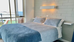 A bedroom with a large bed, patterned bedding, a white brick wall, a small shelf, and a window with a view of the outside. - The Dolphins 7 - A View To Remember (Port Elliot)