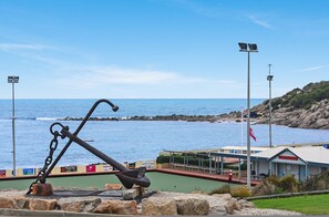 A coastal view with a large anchor sculpture in the foreground, a rocky shoreline, and a building with a sign that reads "South Coast." - The Dolphins 7 - A View To Remember (Port Elliot)