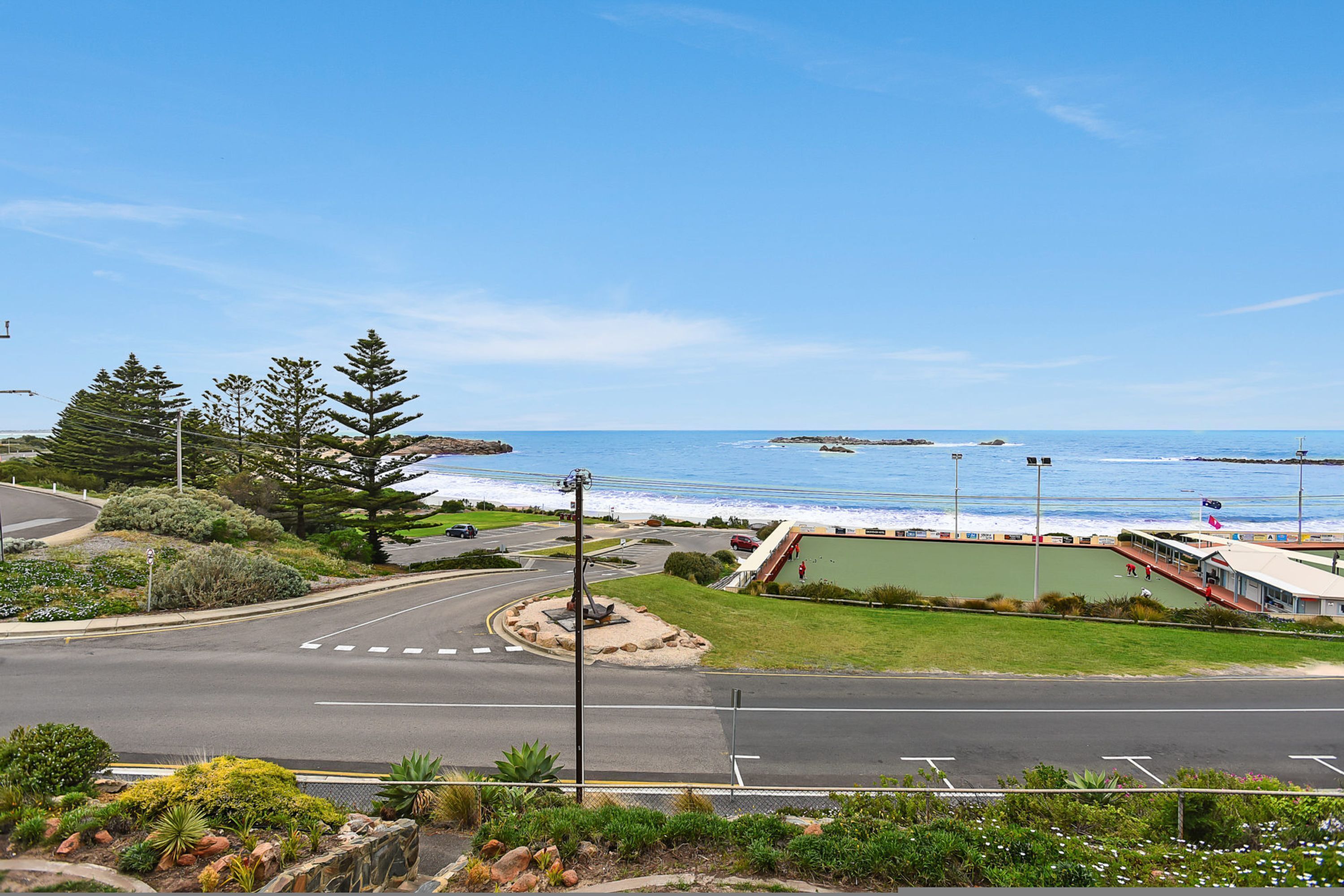 A coastal road with a roundabout, a grassy area, and a building with a green roof near the beach.