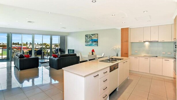 A modern kitchen with white cabinetry, a central island, and a large window offering a view of the outdoors. - Gallery Resort Style Penthouse no 13 (Victor Harbor)