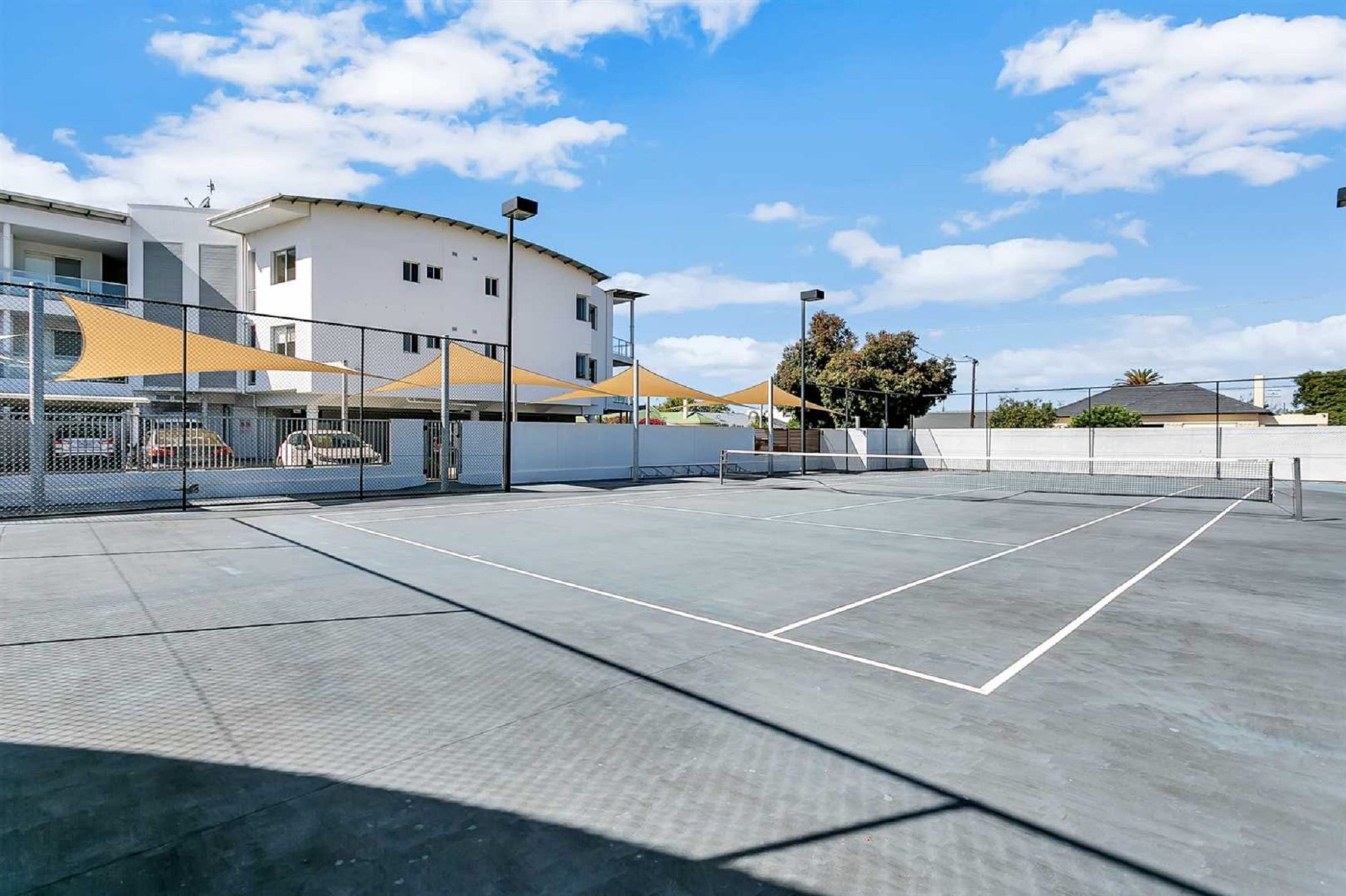 Ein Tennisplatz im Freien mit Netz, umzäunt und mit großen Sonnenschirmen beschattet.