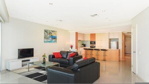 A modern living room with a flat-screen TV, a glass coffee table, and a black leather sofa with red cushions. The kitchen in the background has wooden cabinets and a stainless steel refrigerator. - Gallery Resort Style Penthouse no 13 (Victor Harbor)