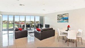 A spacious living area with a large sliding glass door leading to a balcony, a dining area with a white table and chairs, and a painting on the wall. - Gallery Resort Style Penthouse no 13 (Victor Harbor)