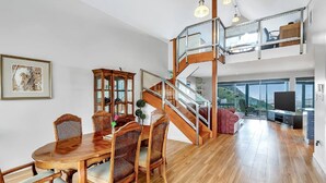A spacious living area with a wooden dining table, chairs, and a staircase leading to an upper level. - The Darling of Marina - A Hilltop and Sea Getaway (Wirrina Cove)