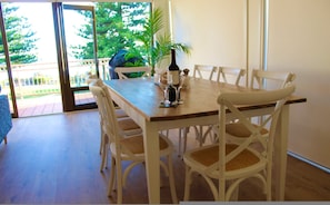 A dining area with a wooden table and white chairs, a bottle of wine, and glasses on the table. - Vista Victor Harbor Beachfront Apartment no 4 (Victor Harbor)