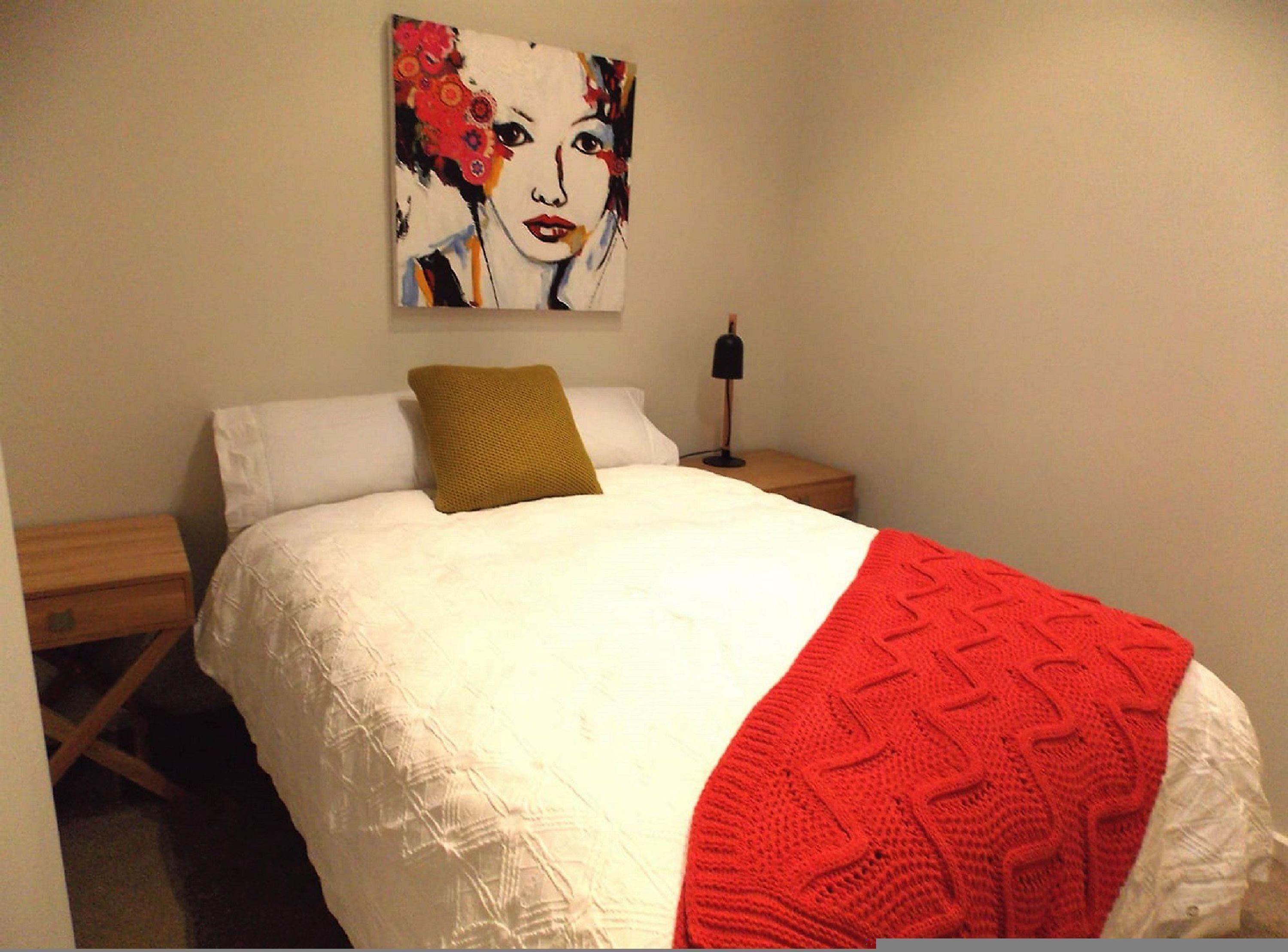 A neatly made bed with a white comforter and a red blanket, a mustard-colored pillow, and a painting on the wall.
