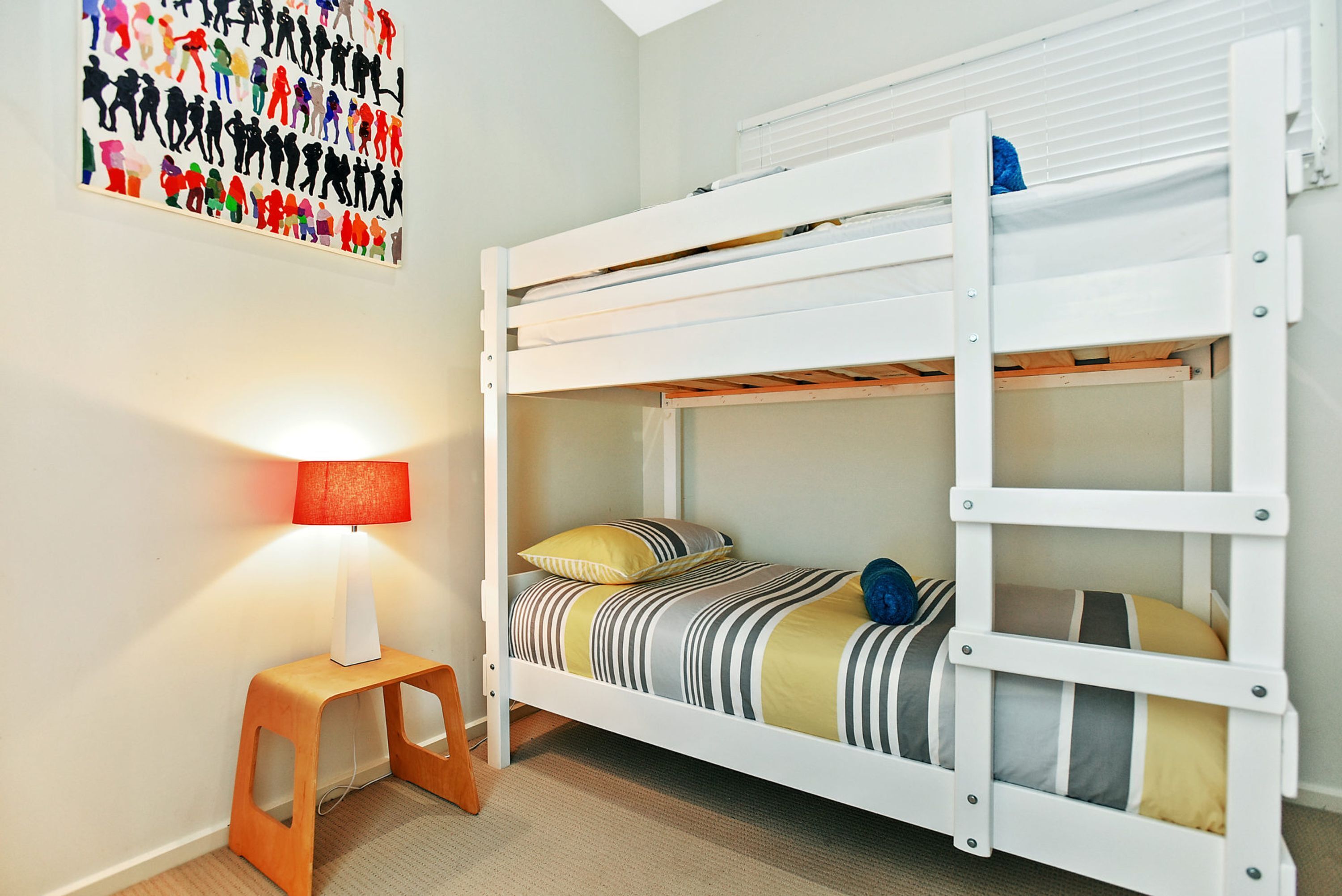 A bunk bed with a striped bedspread and pillows, a small wooden stool, and a wall-mounted lamp.
