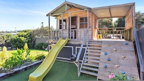 A wooden playhouse with a slide and climbing wall, surrounded by a garden.