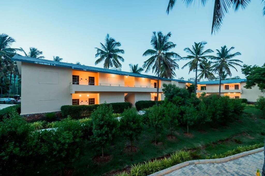 A hotel with a modern design, featuring a flat roof and multiple balconies. The building is surrounded by lush greenery and palm trees, indicating a tropical setting.