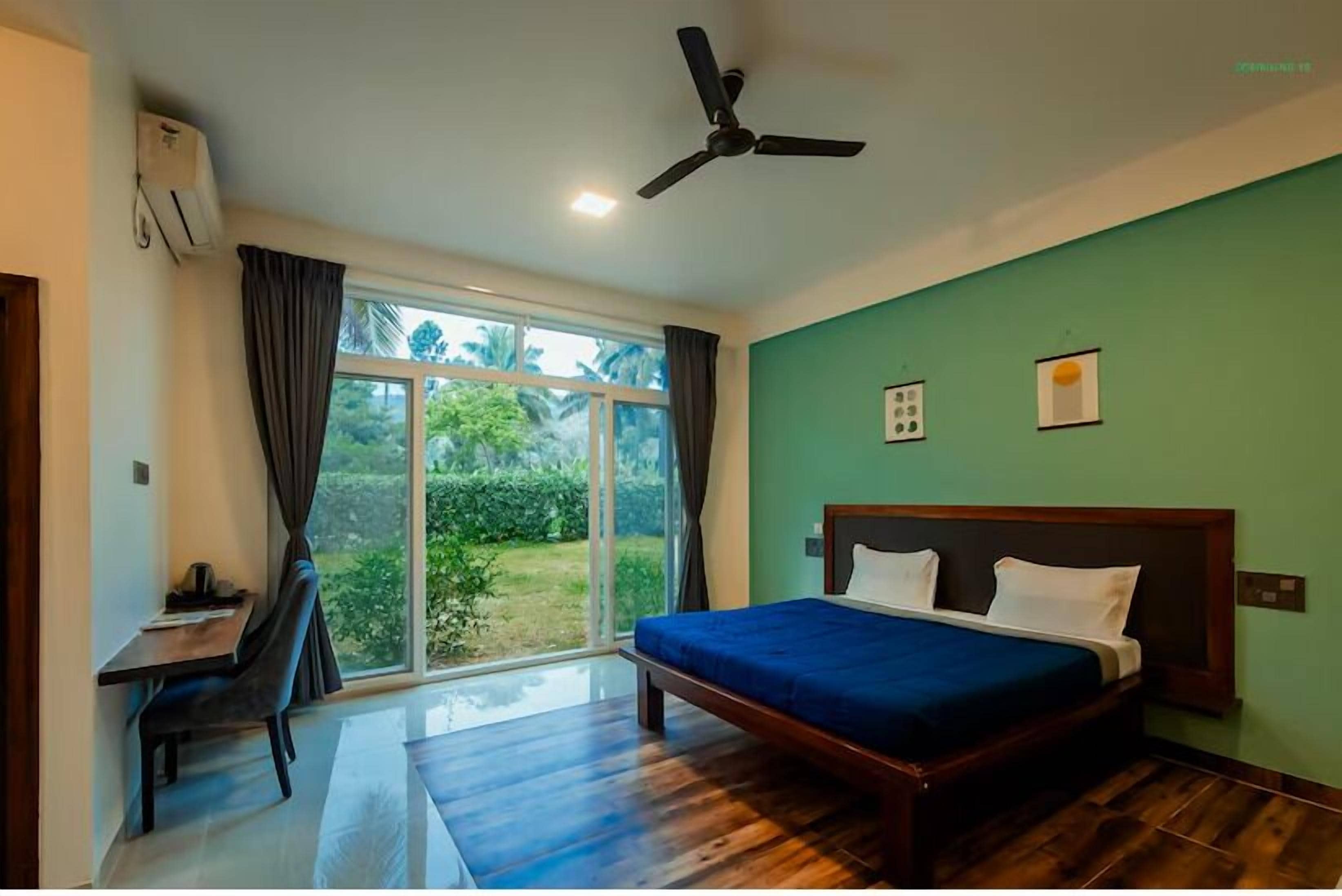 A bedroom with a bed, a desk, and a chair. There is a window with curtains, a ceiling fan, and a view of greenery outside.