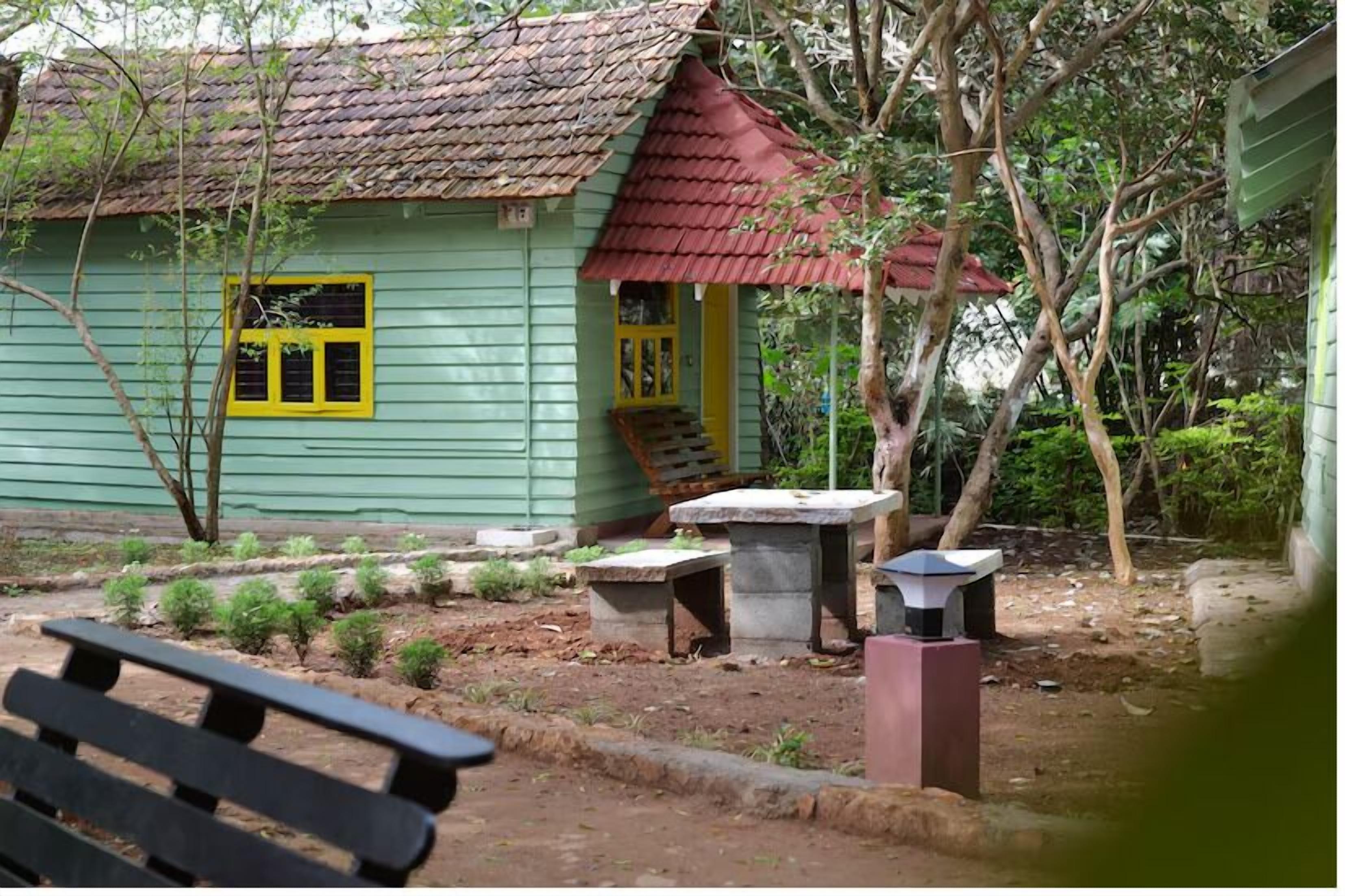 A small, green wooden house with a red roof, surrounded by trees and a small garden.